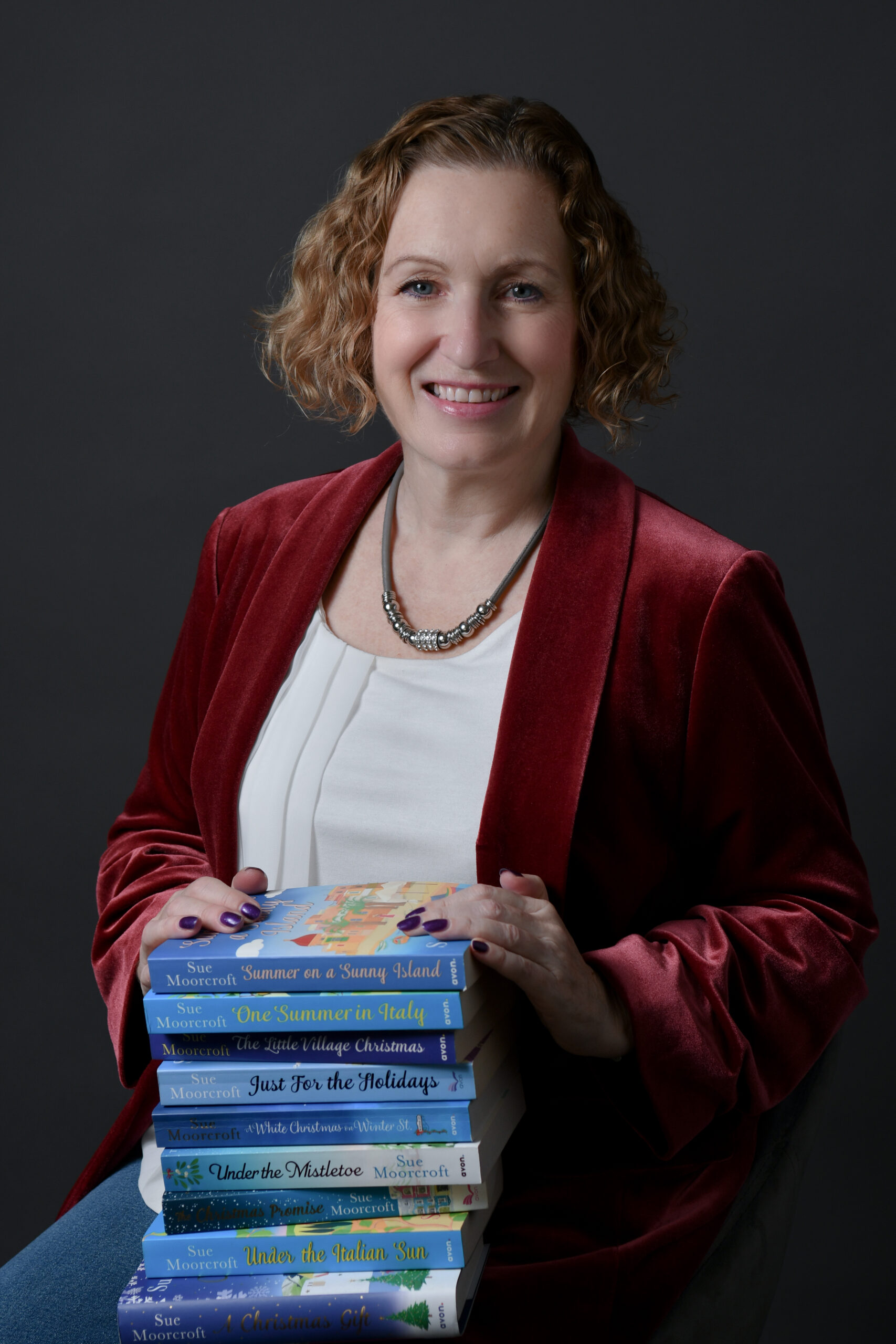 Image shows Sue Moorcroft in red blazer with books